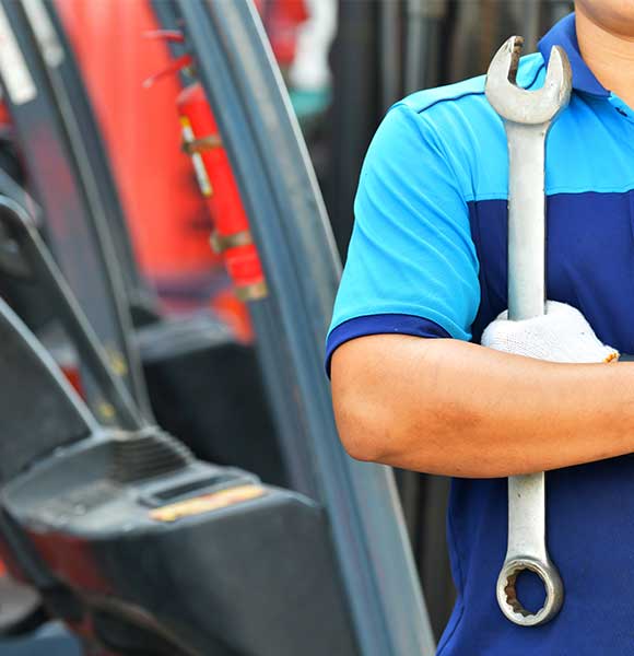 A truck technician wearing a dark blue uniform stands holding a wrench against a blurry forklift background.
